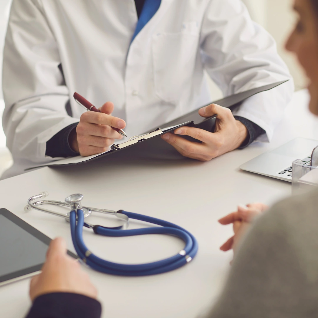 A healthcare professional in a white coat is seated at a table, reviewing patient information on a clipboard while taking notes with a pen. A patient, partially visible, listens attentively. A stethoscope and a tablet are placed on the table, along with a laptop in the background. This image represents a doctor-patient consultation, emphasizing communication and care in a medical setting.