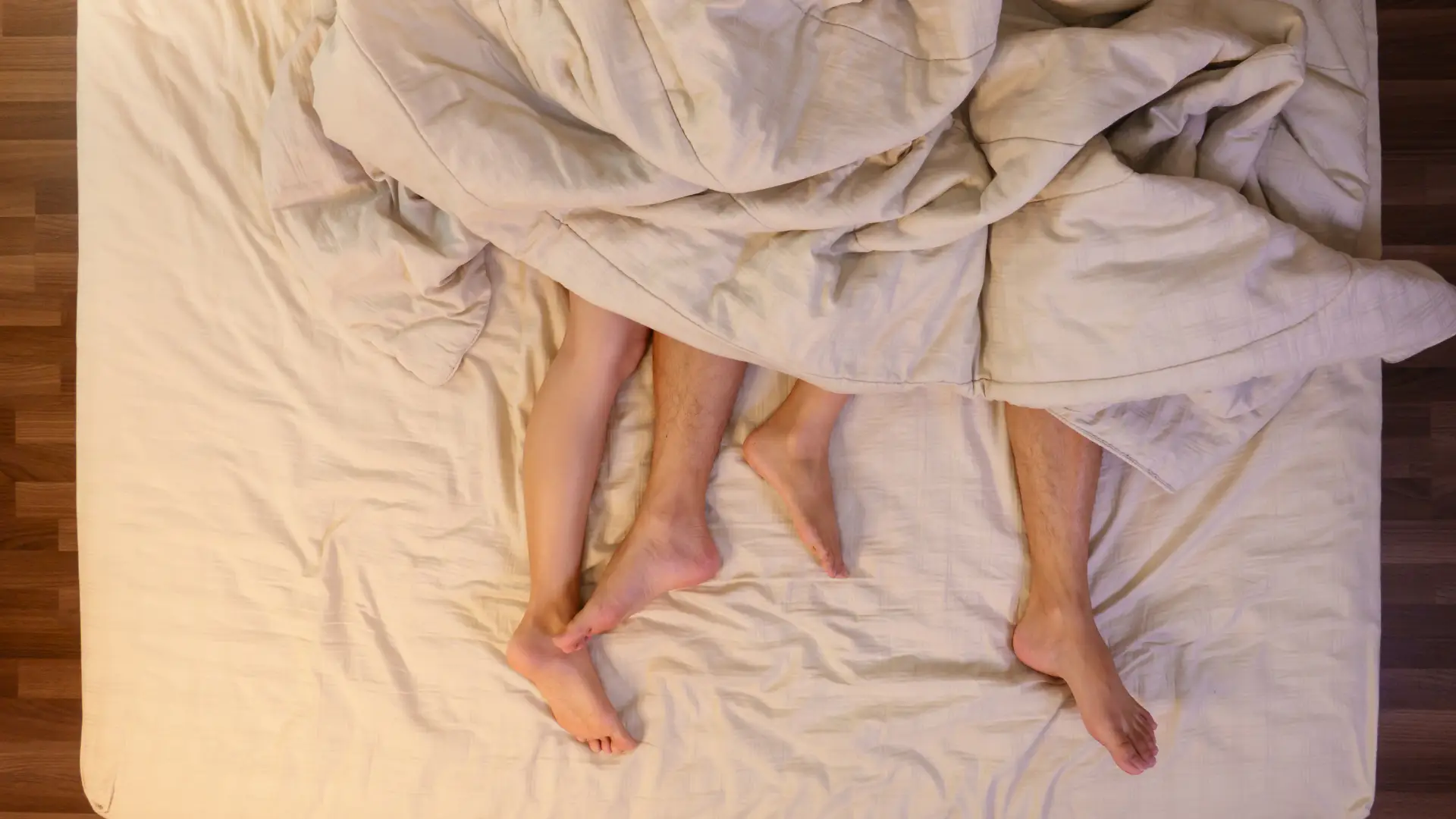 Couple's feet intertwined under a soft, beige blanket on a cozy bed. The image captures a sense of intimacy and comfort, highlighting the connection between the two individuals. The warm tones of the bedding and the gentle arrangement of their legs evoke a peaceful atmosphere, ideal for discussions about relationships, love, or home decor. Perfect for content related to romance, couple activities, or bedroom aesthetics.